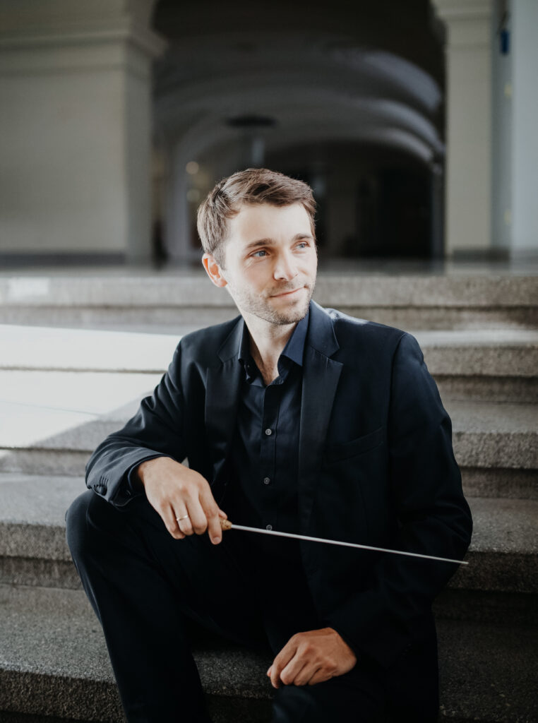 Foto des Dirigenten Tobias Löbner auf einer Treppe sitzend mit Taktstock in der Hand.
